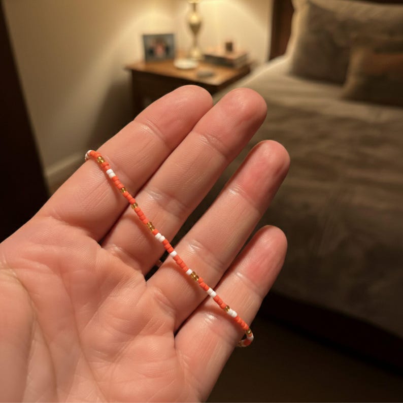 Puede incluir: Una pulsera de cuentas con cuentas de color coral, blancas y doradas. La pulsera se sostiene en una mano, destacando el patr&oacute;n y los colores. Las peque&ntilde;as cuentas est&aacute;n espaciadas uniformemente. Una joya sencilla y elegante.