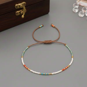 Puede incluir: Pulsera ajustable con un patr&oacute;n de peque&ntilde;as cuentas de colores. La pulsera tiene un cord&oacute;n marr&oacute;n y est&aacute; adornada con cuentas blancas, turquesas, rojas y doradas. La pulsera est&aacute; sobre una superficie gris.
