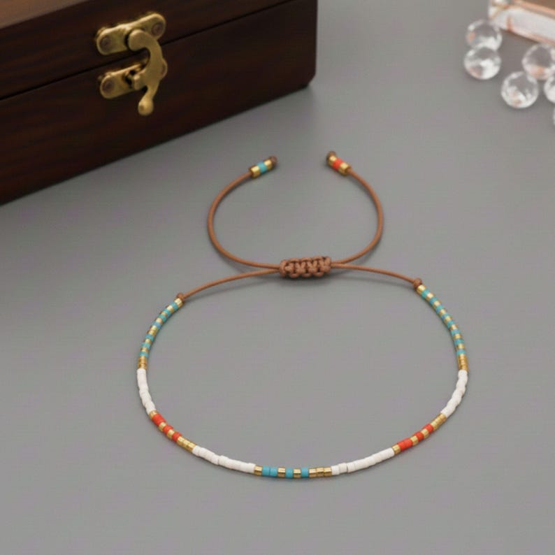 Puede incluir: Pulsera ajustable con un patr&oacute;n de peque&ntilde;as cuentas de colores. La pulsera tiene un cord&oacute;n marr&oacute;n y est&aacute; adornada con cuentas blancas, turquesas, rojas y doradas. La pulsera est&aacute; sobre una superficie gris.