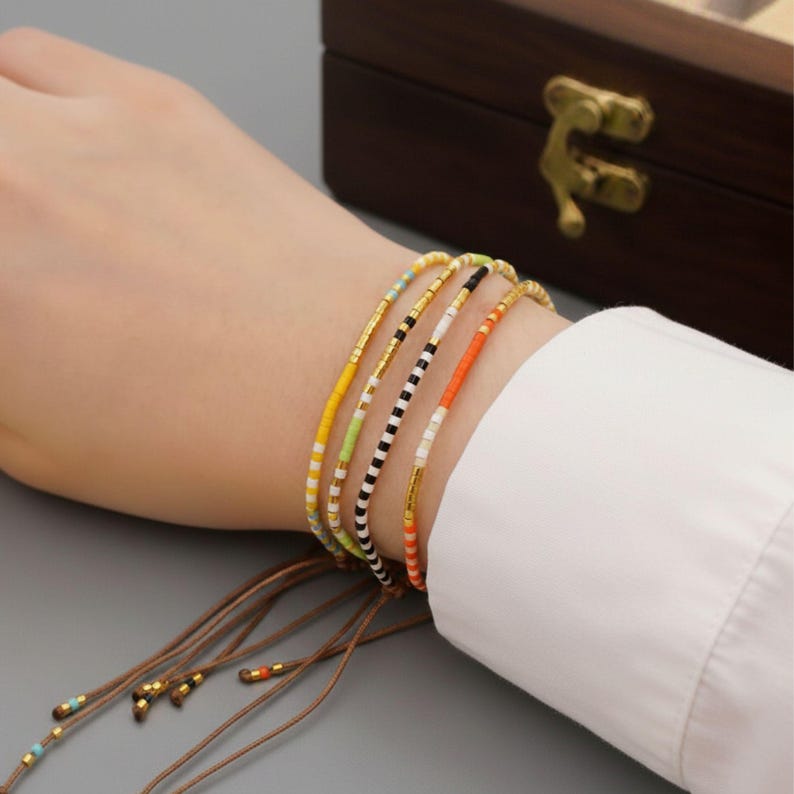 Puede incluir: Una colecci&oacute;n de pulseras de cuentas coloridas con detalles dorados. Las pulseras presentan varios patrones, incluyendo rayas y colores lisos como amarillo, naranja y negro. Las pulseras est&aacute;n en una mu&ntilde;eca, con una cuerda marr&oacute;n y una caja de madera en el fondo.