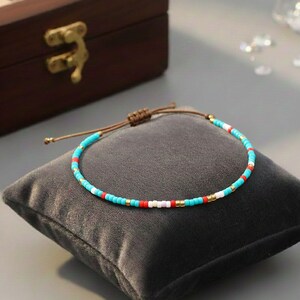 Puede incluir: Una pulsera de cuentas con cuentas turquesas, rojas, blancas y doradas. La pulsera est&aacute; sobre un coj&iacute;n de terciopelo gris. Tiene un cierre de cord&oacute;n marr&oacute;n. Una caja de madera est&aacute; en el fondo.