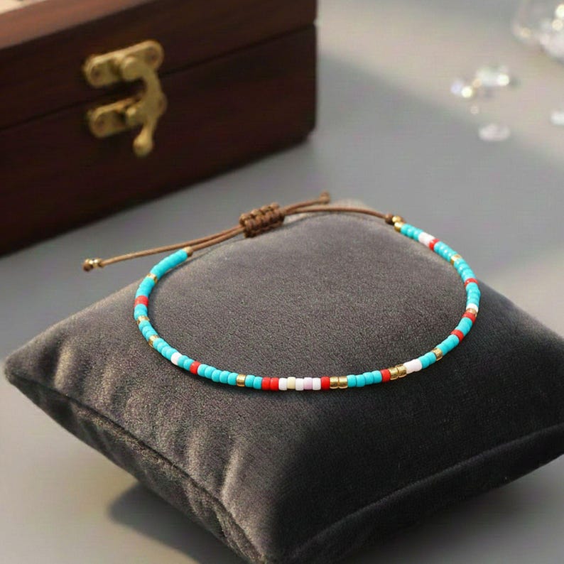 Puede incluir: Una pulsera de cuentas con cuentas turquesas, rojas, blancas y doradas. La pulsera est&aacute; sobre un coj&iacute;n de terciopelo gris. Tiene un cierre de cord&oacute;n marr&oacute;n. Una caja de madera est&aacute; en el fondo.
