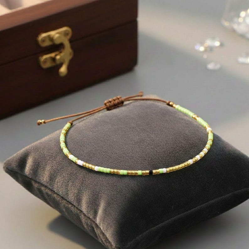 Puede incluir: Una delicada pulsera de cuentas con un patr&oacute;n verde claro, blanco, dorado y negro. La pulsera est&aacute; sobre un coj&iacute;n de terciopelo gris. La pulsera tiene un cord&oacute;n ajustable marr&oacute;n.