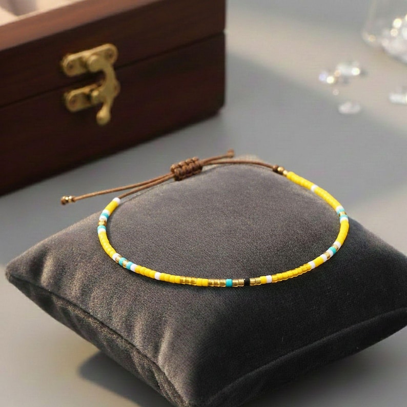 Puede incluir: Una pulsera de cuentas amarillas con detalles blancos, turquesas y negros, sobre un coj&iacute;n de terciopelo gris. La pulsera tiene un cord&oacute;n ajustable marr&oacute;n. Una caja de madera con un cierre dorado est&aacute; en el fondo.