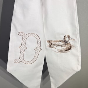 以下が含まれることがあります： 茶色の刺繍が施された白い生地。片側には、格子模様の「D」の文字が刺繍されています。もう一方には、水中で泳ぐアヒルの刺繍が施されています。生地の底は尖っています。