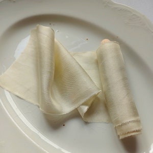 May include: A length of ivory silk ribbon, partly rolled and unrolled, displayed on a white, oval plate. The ribbon has a gentle sheen and a frayed edge. The colour of the ribbon is a soft, creamy hue, suggesting a delicate texture.