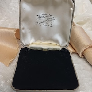 May include: A square jewellery box with a hinged lid, featuring a cream-coloured satin interior lining and a black velvet base. The inside lid is printed with the text: Established 1790, Goldsmiths, Silversmiths & Diamond Merchants, Bracher & Sydenham, Reading.