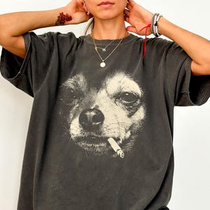 Puede incluir: Una camiseta gris carbón de corte oversize con un gran estampado gráfico de alto contraste que muestra la cara de un perro con un cigarrillo en la boca. La prenda tiene cuello redondo, mangas cortas y un ajuste holgado con acabado desgastado.