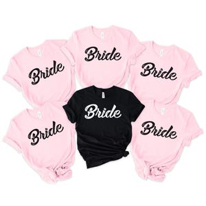 May include: A selection of pink and black t-shirts, each displaying the word "Bride" in a black script font. These short-sleeved shirts are likely made from a soft fabric, suitable for a hen party or bridal group.