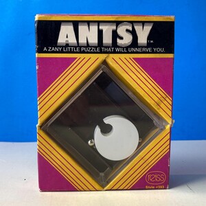 Könnte beinhalten: Vintage "ANTSY" Puzzle-Box mit einem schwarz-weiß-gelben Design. Die Box zeigt den Text "ANTSY" und "A ZANY LITTLE PUZZLE THAT WILL UNNERVE YOU." Das Puzzle selbst ist durch ein transparentes Kunststofffenster sichtbar.