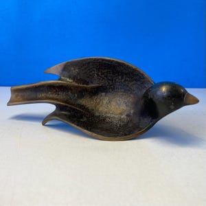 Puede incluir: Un plato con forma de pájaro de color bronce con una superficie texturizada. El pájaro tiene las alas extendidas y un pico puntiagudo. El plato probablemente se utiliza con fines decorativos o como un pequeño recipiente.