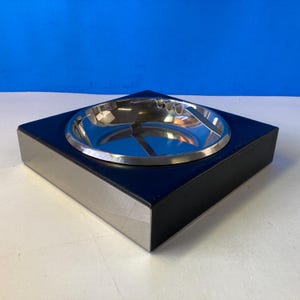 以下が含まれることがあります： 黒とシルバーのベースにステンレス製のボウルがセットされた正方形の灰皿。 灰皿は、すっきりとしたミニマリストな美学を備えたモダンなデザインです。 ボウルは、タバコを置くための溝が付いた湾曲したデザインです。
