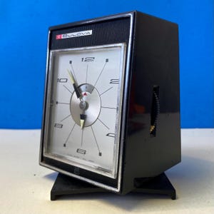 Op de afbeelding: Een vintage zwarte Bulova klok met een zilveren wijzerplaat en zwarte wijzers. De klok heeft een rechthoekige vorm en staat op een zwarte standaard. De wijzerplaat heeft zwarte uurmarkeringen en het Bulova-logo.