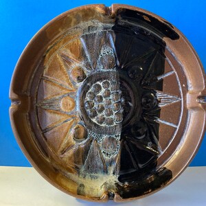 May include: A round ceramic ashtray with a brown and black glaze. The ashtray has a decorative design with a central circle and radiating lines.