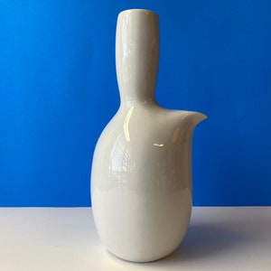 May include: A white ceramic jug with a distinctive, curved design. The jug has a spout on the side and a wide opening at the top.