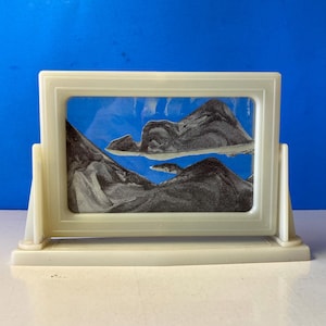 May include: A cream-colored sand art picture frame with a landscape scene. The frame holds a blue background with black and gray sand creating a mountain and lake scene. The frame is rectangular and sits on a stand.