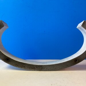 May include: A gray concrete curved shape with a smooth, slightly textured surface. The shape is open on one side and has a rounded edge on the other side. There are several small, round holes along the edge of the shape.