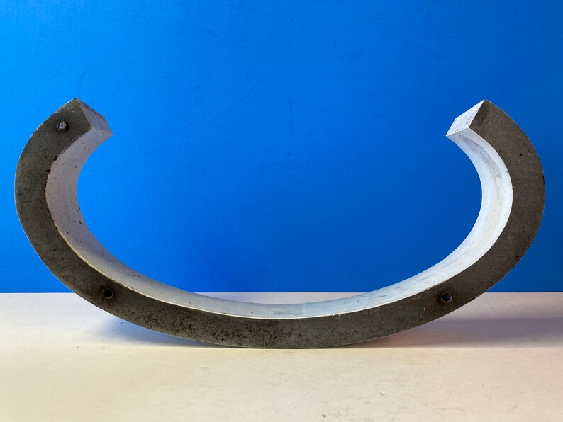 May include: A gray concrete curved shape with a smooth, slightly textured surface. The shape is open on one side and has a rounded edge on the other side. There are several small, round holes along the edge of the shape.