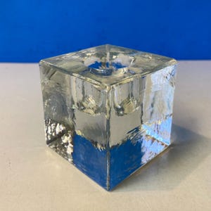Puede incluir: Un portavelas cuadrado de vidrio transparente con una superficie texturizada. El portavelas tiene una hendidura circular en la parte superior para una vela. El diseño en forma de cubo añade un toque moderno a la decoración del hogar.