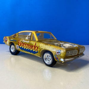 Puede incluir: Un modelo de coche Plymouth Barracuda dorado con letras azules y rojas. El modelo tiene un capó transparente, que revela el motor. El coche tiene logotipos de Goodyear y STP. El texto "426 HEMI" es visible.