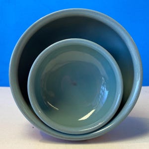 May include: Two nested, light blue ceramic bowls. The larger bowl surrounds a smaller bowl, both with a glossy finish. The bowls are set against a blue background, creating a contrast that highlights their shape and color.