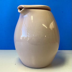 May include: A pale pink ceramic pitcher with a rounded body and a spout. The pitcher has a lid with a rounded top.