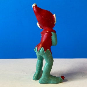 Op de afbeelding: Een keramiek elfenbeeldje met een rode muts en een groene broek. De elf kijkt omhoog en heeft een speelse uitdrukking.