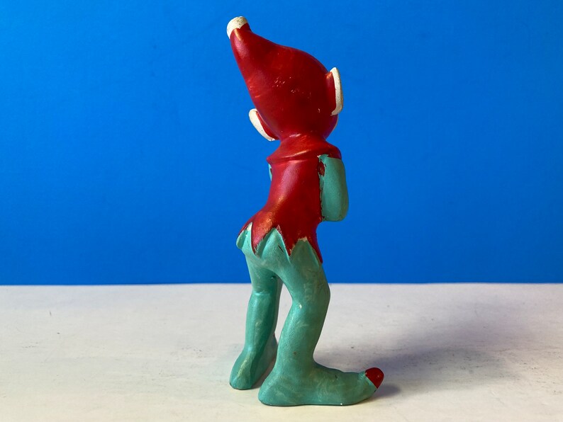 Op de afbeelding: Een keramiek elfenbeeldje met een rode muts en een groene broek. De elf kijkt omhoog en heeft een speelse uitdrukking.