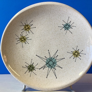 Op de afbeelding: Een wit keramiek dinerbord met een gespikkelde glazuur en een mid-century modern design met zwarte lijnen en gele en blauwe accenten.