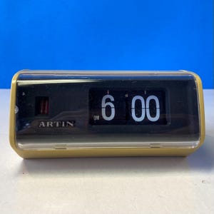 May include: A vintage flip clock with a yellow frame and a clear plastic cover. The clock displays "6:00 PM" in white numerals against a black background. The brand name "ARTIN" is visible on the front.