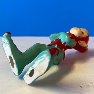 Op de afbeelding: Een klein keramiek beeldje van een elf die op zijn rug ligt. De elf draagt een rode muts en een blauw en rood pakje. De voeten van de elf zijn zichtbaar en zijn wit geschilderd.