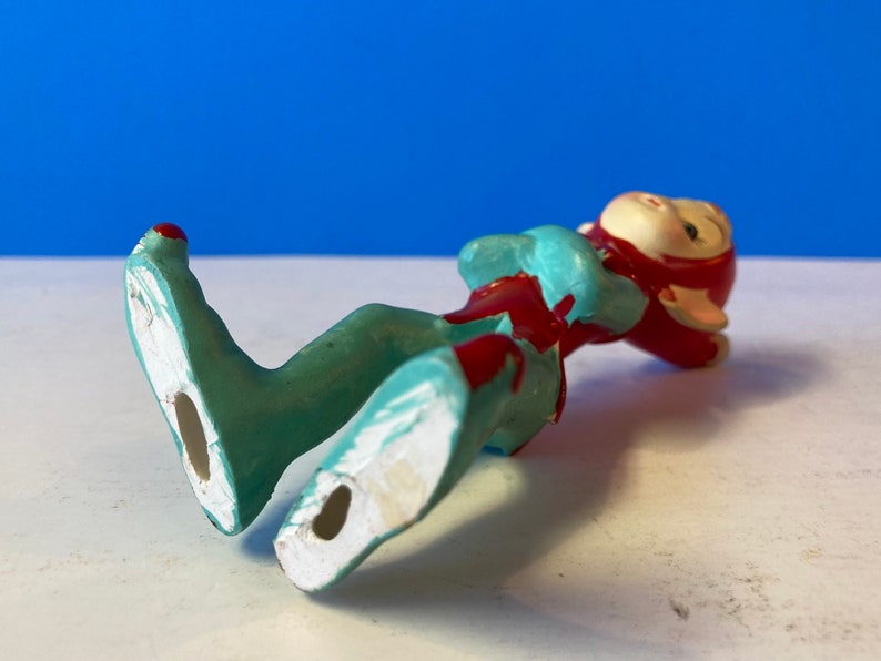 Op de afbeelding: Een klein keramiek beeldje van een elf die op zijn rug ligt. De elf draagt een rode muts en een blauw en rood pakje. De voeten van de elf zijn zichtbaar en zijn wit geschilderd.