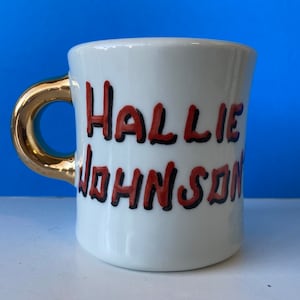May include: White ceramic mug with a gold handle. The mug features the name "Hallie Johnson" in red letters with black outlines. The mug is set against a blue background.