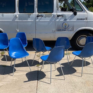 May include: A collection of eight blue plastic chairs with silver metal legs. The chairs are arranged outdoors in front of a white van. The chairs have a mid-century modern design.