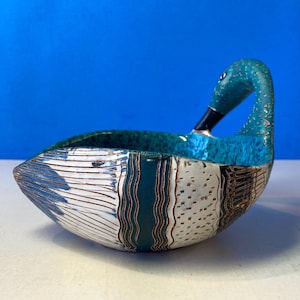 May include: A ceramic duck-shaped bowl with a teal and white exterior. The duck's head and neck are teal, with a black beak. The bowl's body features vertical stripes and dotted patterns, with brown and teal accents.