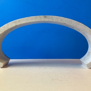 May include: A white concrete arch sculpture. The arch is curved and has a smooth, slightly textured surface.