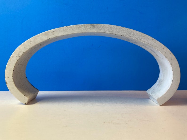 May include: A white concrete arch sculpture. The arch is curved and has a smooth, slightly textured surface.