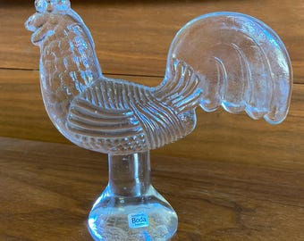 May include: A clear glass rooster figurine with a detailed design. The rooster has a comb, wattle, and tail feathers. The figurine is mounted on a rounded base. The base has a blue and white sticker that reads "Handmade Boda Sweden".
