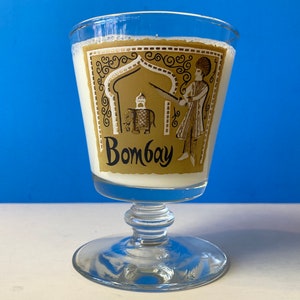 May include: A clear glass goblet with a gold and white design featuring an elephant and a man in traditional clothing. The goblet has a stemmed base and says "Bombay" on the front.