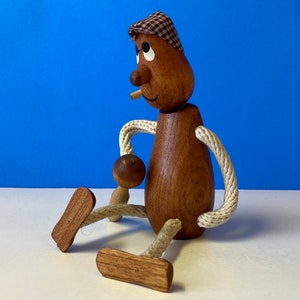 May include: A wooden figurine of a person with a plaid hat, brown eyes, and a painted-on smile. The figurine has rope arms and legs, and is sitting with one leg extended.