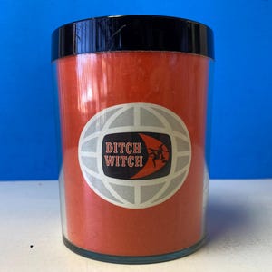 Puede incluir: Un tarro de vidrio rojo con tapa negra y una etiqueta blanca que dice "Ditch Witch" con un gráfico de globo terráqueo.