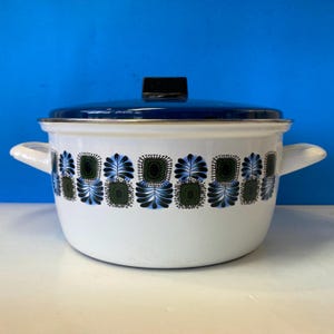May include: A white enamel stockpot with a dark blue lid and two side handles. The exterior features a repeating horizontal pattern of stylised dark green and blue floral motifs encircling the body of the pot, with a metallic rim at the top.