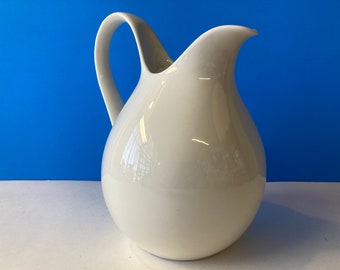 May include: A white ceramic pitcher with a rounded body and a curved spout. The pitcher has a smooth, glossy finish.
