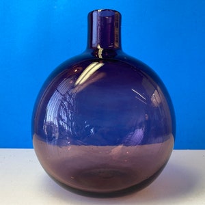 May include: A round, dark blue glass bottle with a short neck. The bottle has a bulbous shape and a smooth surface. The glass reflects light, creating highlights and shadows. The bottle is likely used for decorative purposes or as a vase.