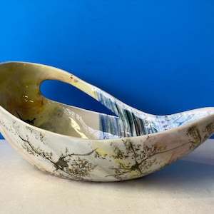 May include: A white ceramic bowl with a unique, abstract design featuring splashes of yellow, blue, green, and brown. The bowl has a handle on one side and a slightly pointed end on the other.
