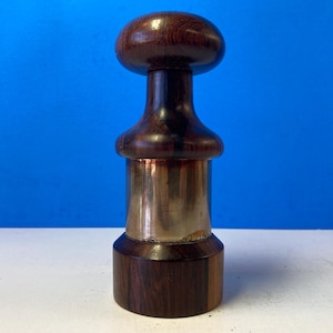 Może przedstawiać: Drewniany i mosiężny korek do butelki z zaokrąglonym wierzchołkiem i cylindryczną podstawą. Wierzchołek jest wykonany z ciemnobrązowego drewna z widocznym usłojeniem. Podstawa jest wykonana z ciemnobrązowego drewna z mosiężnym paskiem wokół środka.