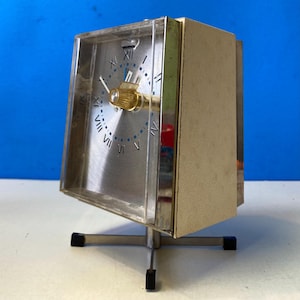 May include: A vintage silver and gold-toned clock with a clear plastic cover. The clock face has Roman numerals and blue accents. It stands on a silver metal base with black feet. The clock is a retro style.