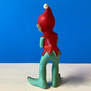 Op de afbeelding: Een rood en groen keramiek elfenbeeldje met een punthoed. De elf staat met gekruiste armen en kijkt naar de zijkant.