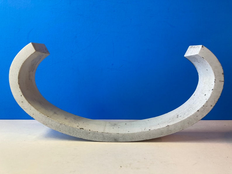May include: A concrete half-circle sculpture with a smooth, gray surface. The sculpture is curved and has a rounded edge.