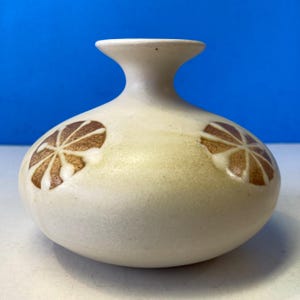 May include: A small, off-white ceramic vase with a rounded body and a narrow neck. Two brown floral designs are on the sides. The vase has a matte finish and a simple, elegant design, suitable for displaying small flowers or as a decorative piece.
